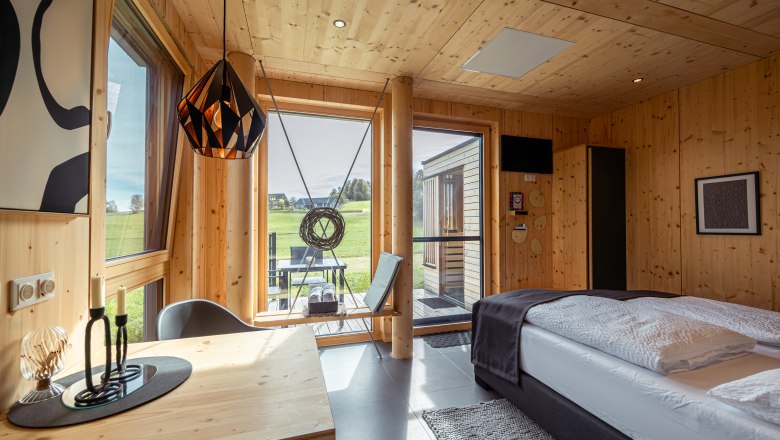 Innenansicht eines modernen Holzhauses mit Bett, Tisch und gro&szlig;en Fenstern mit Blick ins Freie.