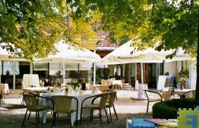 Ein gemütlicher Gastgarten mit Tischen und Stühlen unter Sonnenschirmen, umgeben von Bäumen.