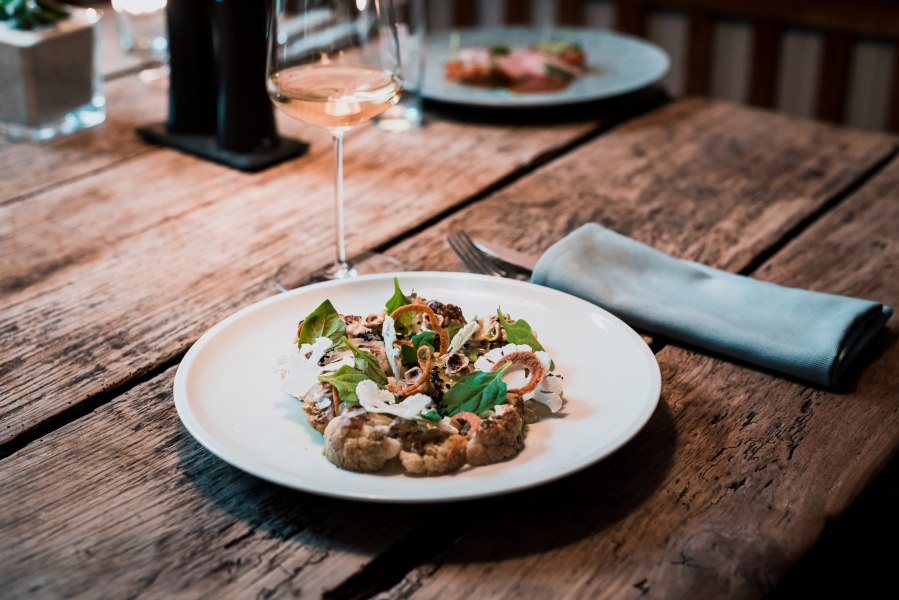 Ein Teller mit geschmortem Karfiol, Miso Spinat und eingelegtem Sanddorn auf einem Holztisch, daneben ein Glas Wei&szlig;wein.