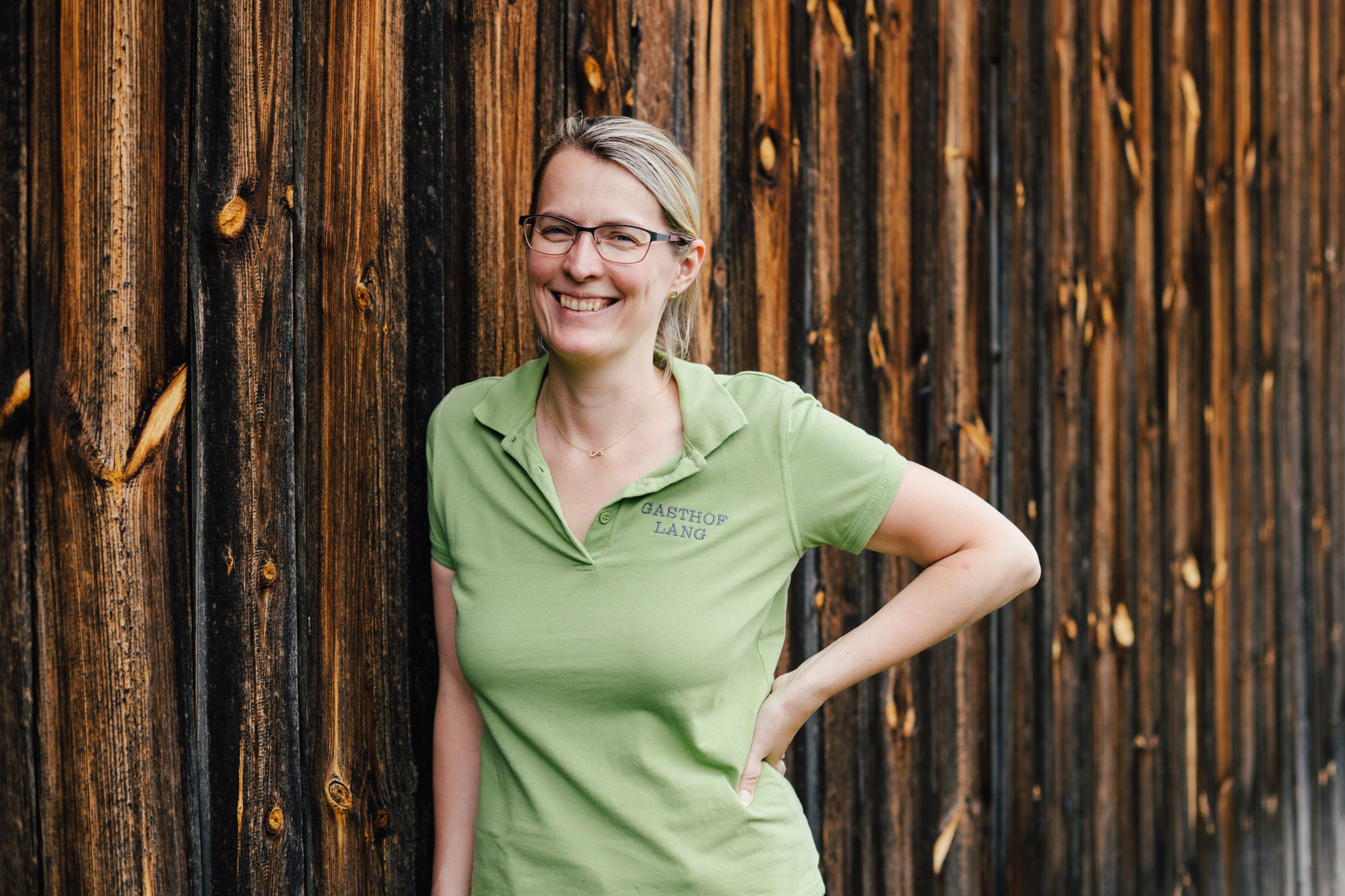 Eine Frau in einem grünen Polohemd lehnt lächelnd an einer Holzwand.