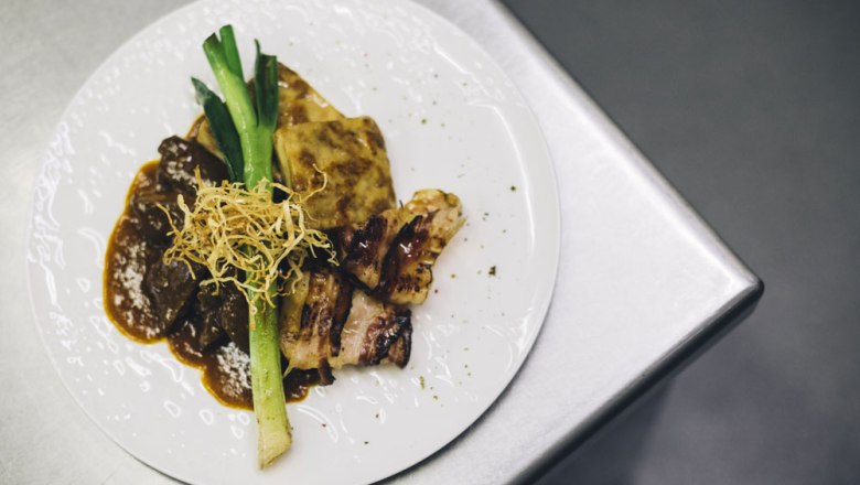 Ein Teller mit Rostbraten, Kartoffelgratin, Lauch und knusprigen Zwiebeln auf einer Metalloberfl&auml;che.
