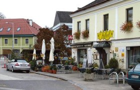 Außenansicht von einem Gasthaus an einem Belebten Platz mit Bäumen, Sonnenschirmen, Sesseln uvm.