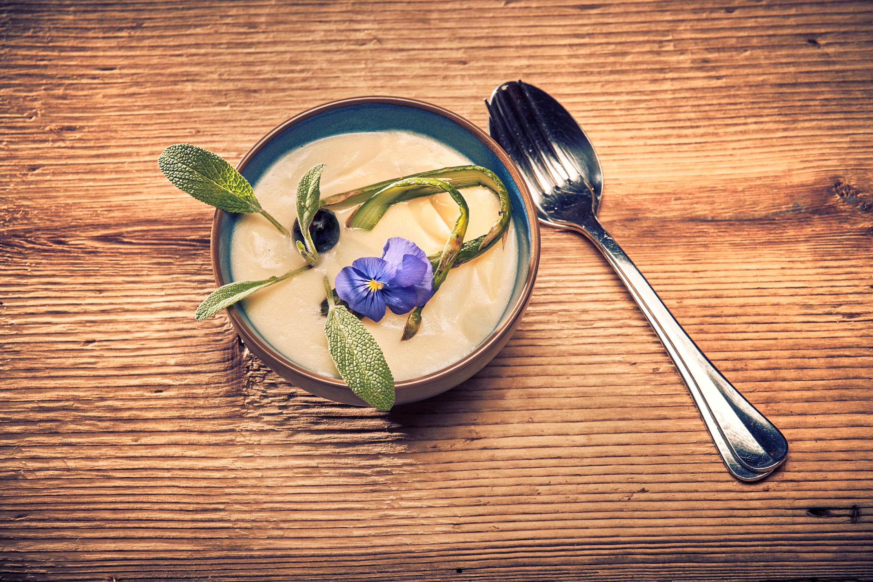Spargelcremesuppe Suppe mit Blüte und Kräutern auf Holztisch, Löffel daneben.
