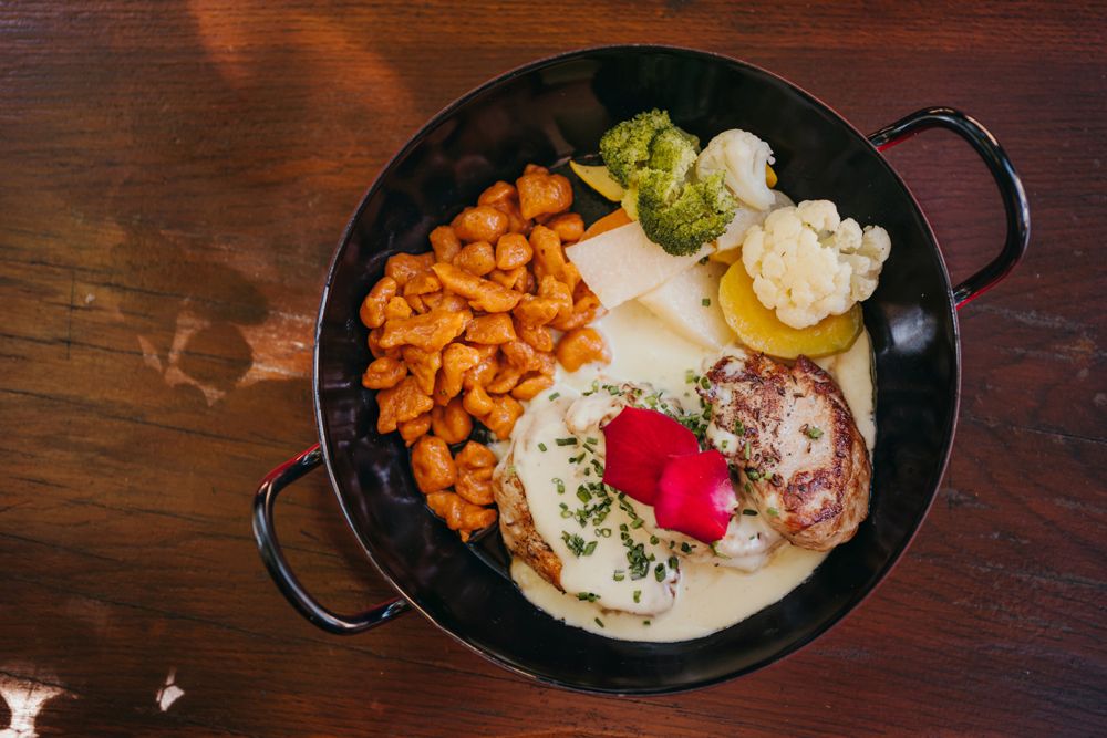 Schweinsmedaillons mit Käsesauce, roten Spätzle und Gartengemüse in einer schwarzen Pfanne.