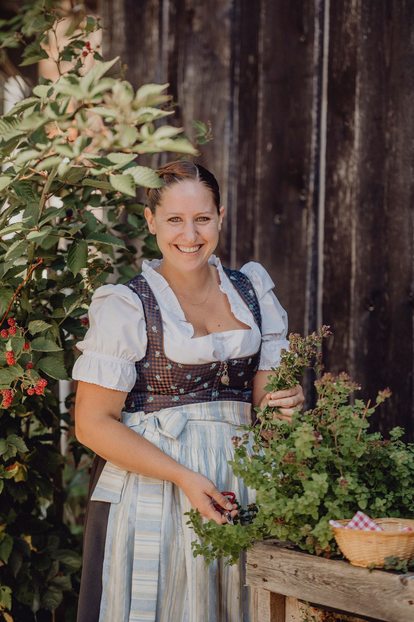 Eine Frau in traditioneller Tracht steht lächelnd neben einem Strauch und hält Kräuter in der Hand.