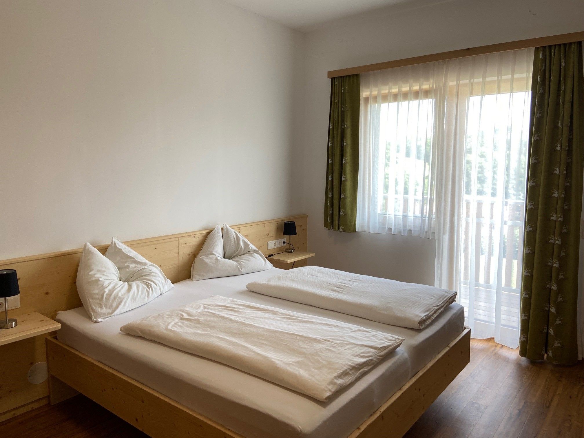 Ein helles Schlafzimmer mit einem Doppelbett, weißen Bettdecken und Kissen. Ein Fenster mit grünen Vorhängen lässt Tageslicht herein.