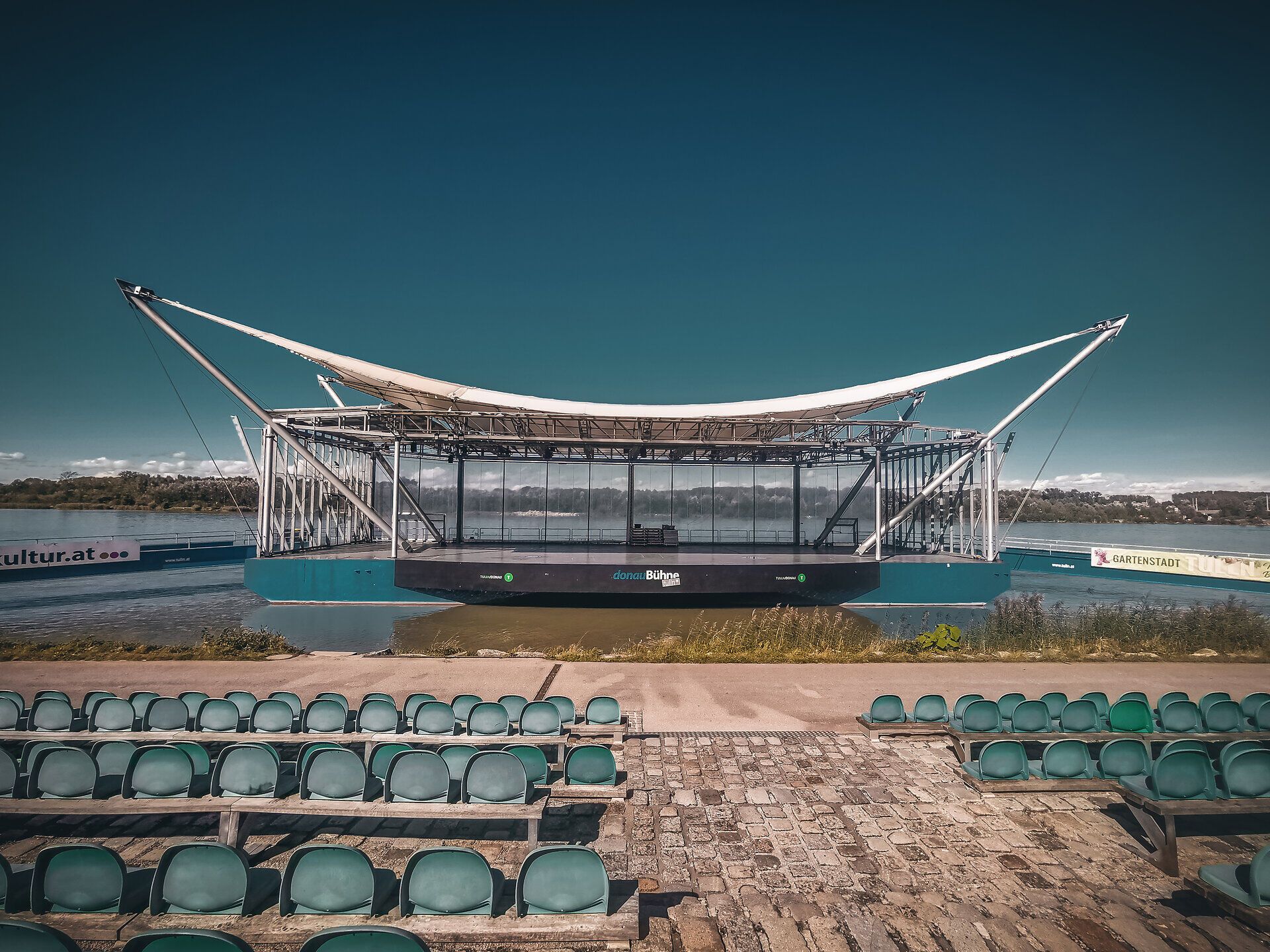 Die sanften Wellen der Donau spiegeln die warmen Farben des Herbstes wider, während die Sitzreihen am Ufer auf unvergessliche Veranstaltungen warten. Ein Ort der Ruhe und Inspiration, wo die Natur und Kultur harmonisch verschmelzen.