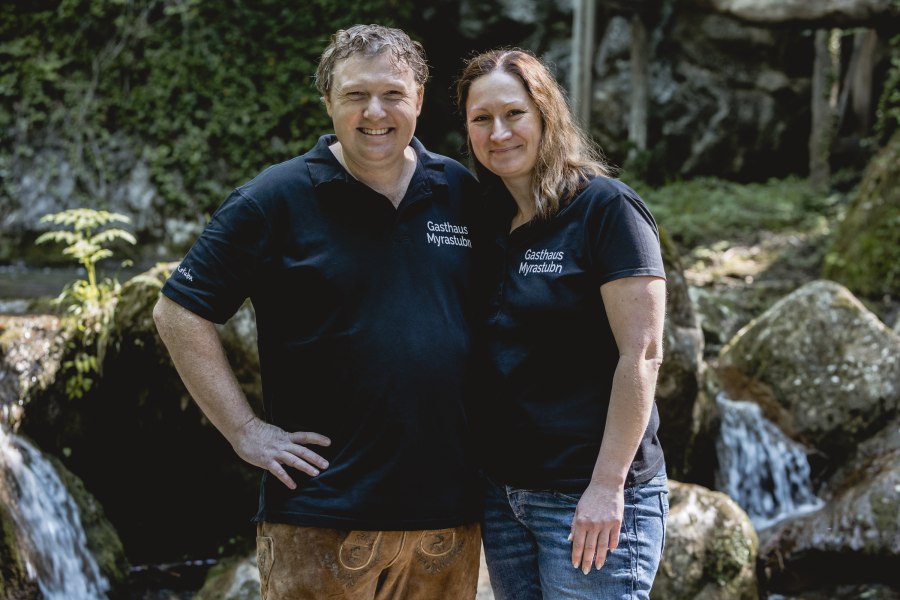 Ein Mann und eine Frau in schwarzen T-Shirts mit der Aufschrift 'Gasthaus Myrastubn' stehen lächelnd vor einem Wasserfall.