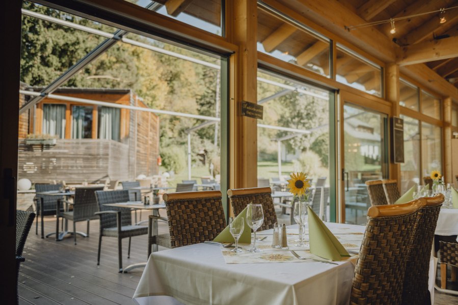 Ein Restaurant mit Wintergarten und Terrasse, gedeckte Tische mit Sonnenblumen, Blick auf gr&uuml;ne Umgebung.