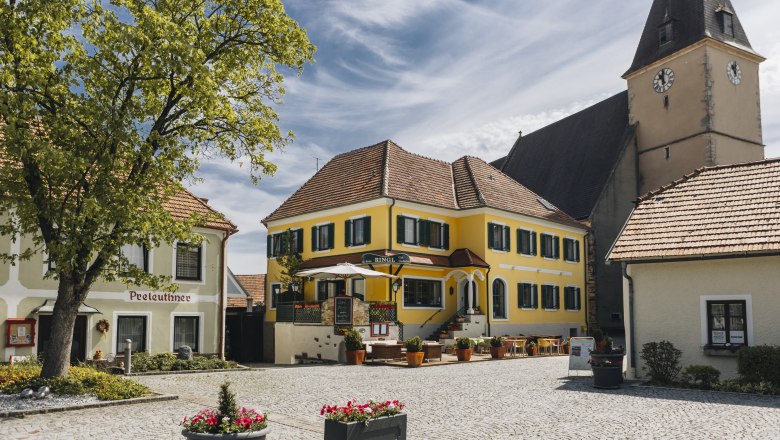 Idyllischer Dorfplatz mit Gasthaus und Terrasse und ein Kirchturm im Hintergrund.