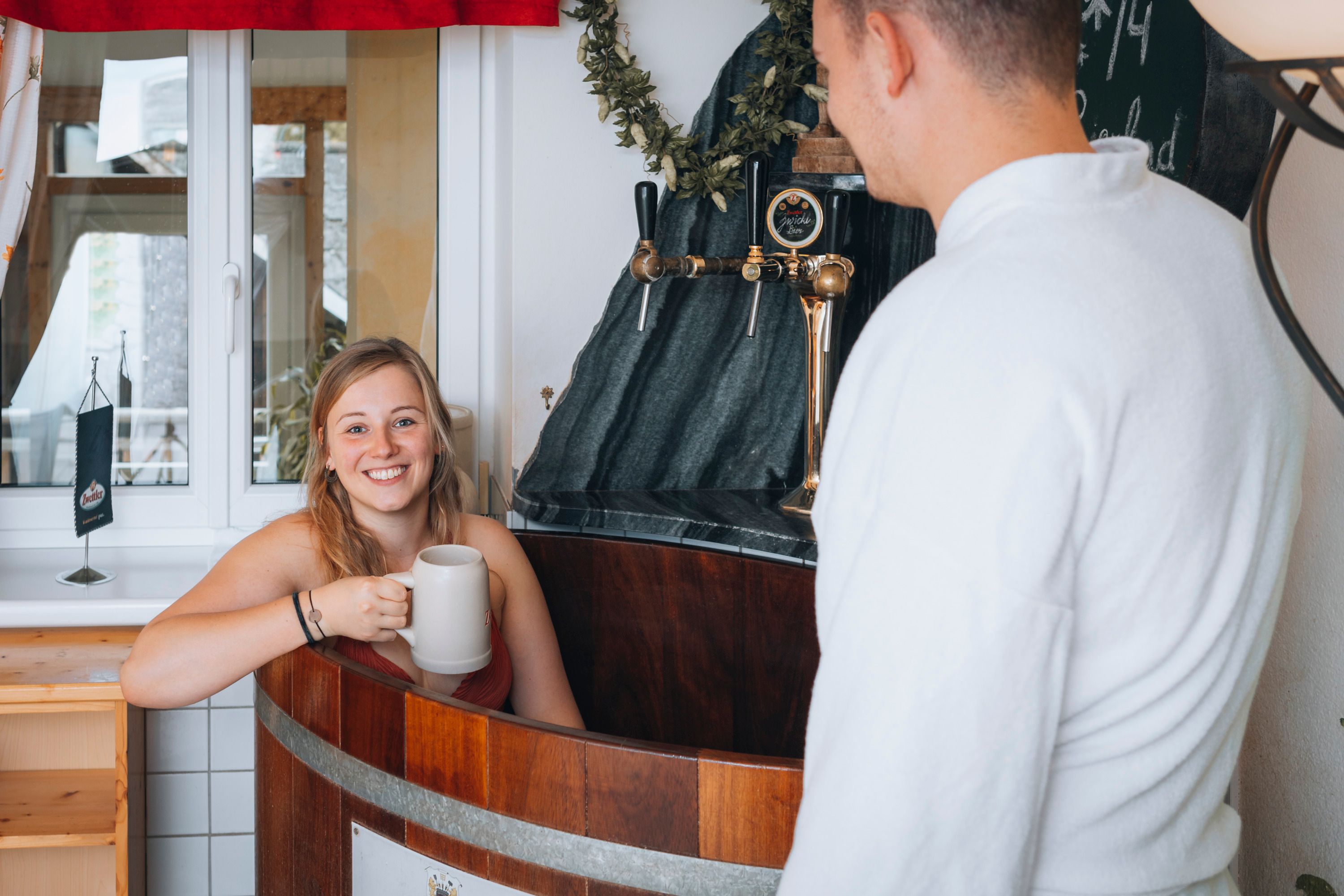 Frau in einem Bierbad mit Krug, Mann im Bademantel daneben.