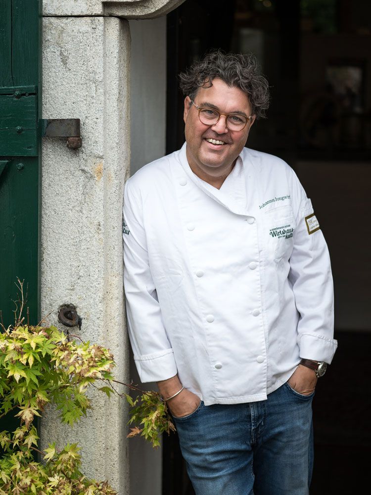 Mann in weißer Kochjacke lehnt lächelnd an einer Tür.