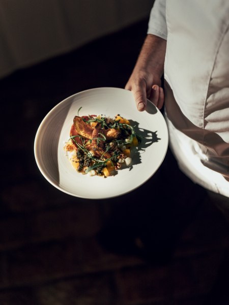 Ein Teller mit geröstetem Hokkaido-Kürbis, Salbeipesto und Schafkäse, gehalten von einer Person in weißer Kleidung.