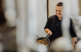 Eine Köchin in schwarzer Uniform hält eine Pfanne mit gebratenem Gemüse und lächelt.