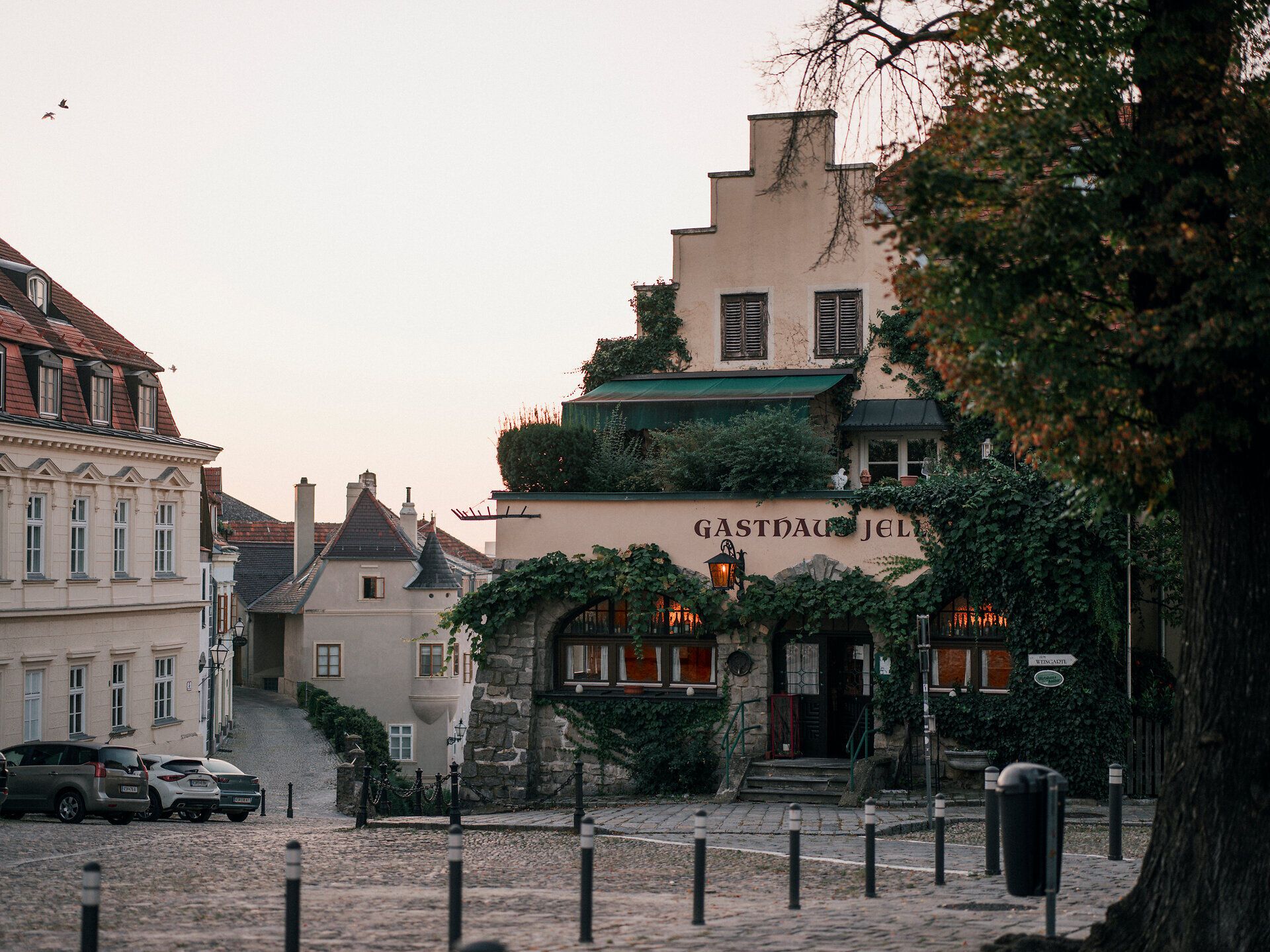 In der sanften Dämmerung erstrahlt das Gasthaus Jell, umgeben von üppigem Grün und historischen Gebäuden. Die gepflasterten Straßen laden zu einem gemütlichen Spaziergang ein, während die warmen Lichter des Wirtshauses eine einladende Atmosphäre schaffen.