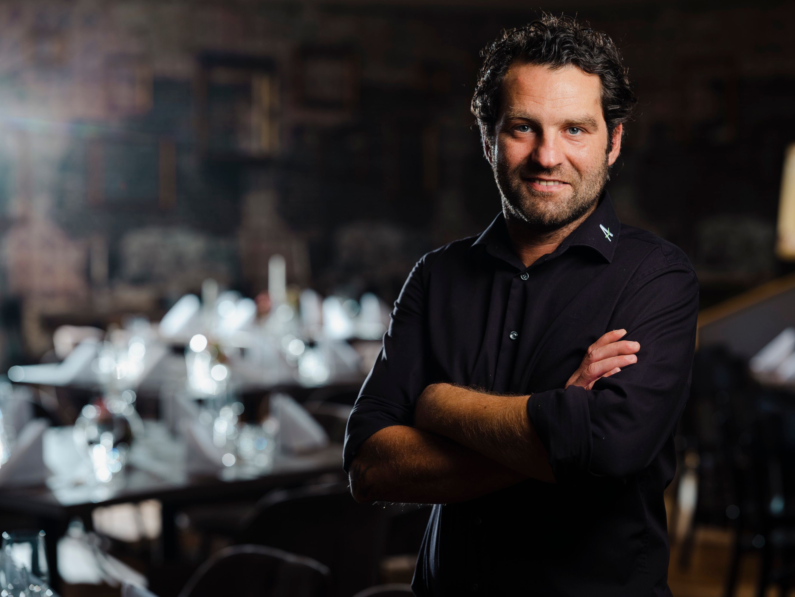 Mann in schwarzem Hemd steht in elegantem Restaurant, Tische im Hintergrund.