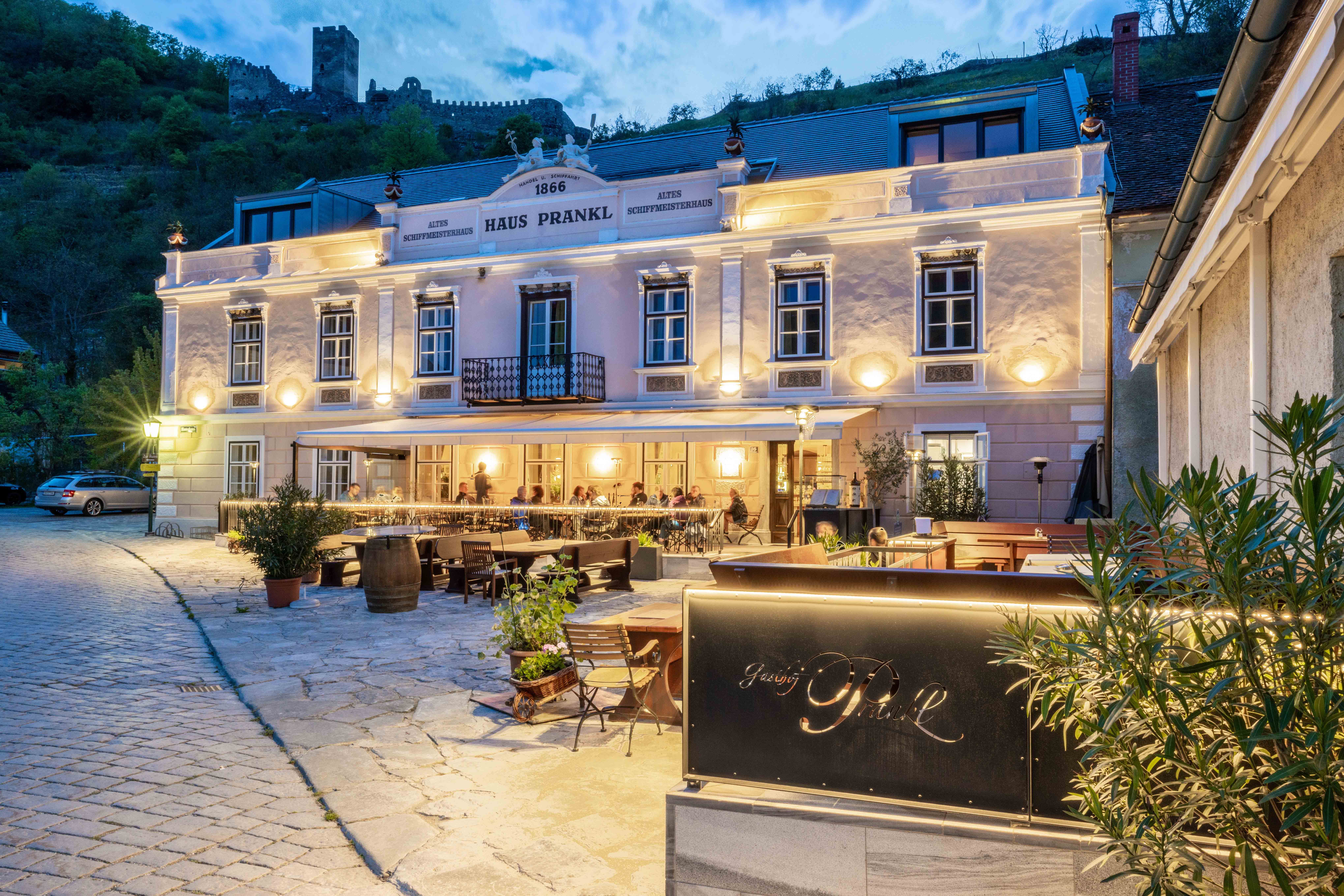 Außenansicht des Gasthofs Prankl bei Abenddämmerung mit beleuchteter Terrasse und Burg im Hintergrund.