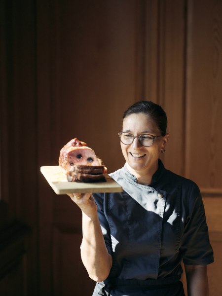 Eine Frau in Kochkleidung h&auml;lt ein Holzbrett mit einem aufgeschnittenen Fleischgericht.
