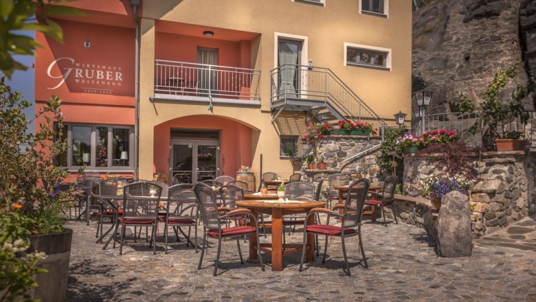 Außenbereich eines Restaurants mit Tischen und Stühlen auf einem gepflasterten Hof, umgeben von Blumen und Pflanzen.