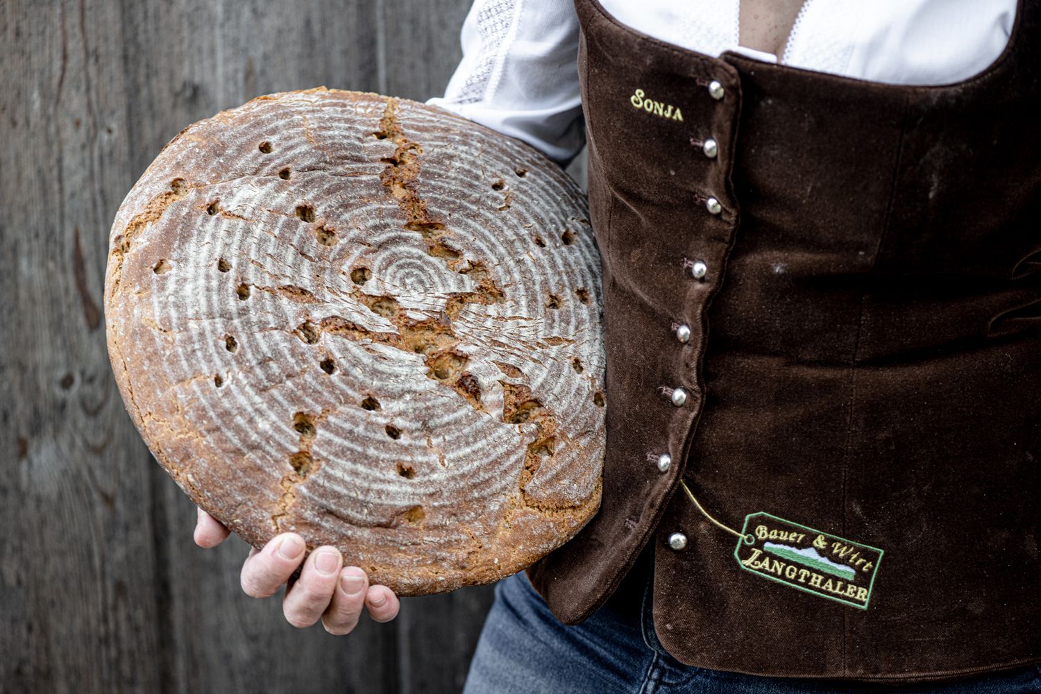 Person hält rundes, selbstgebackenes Brot vor rustikalem Hintergrund.
