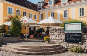 Gelbes historisches Gebäude mit Terrasse und Sonnenschirmen, umgeben von Pflanzen.