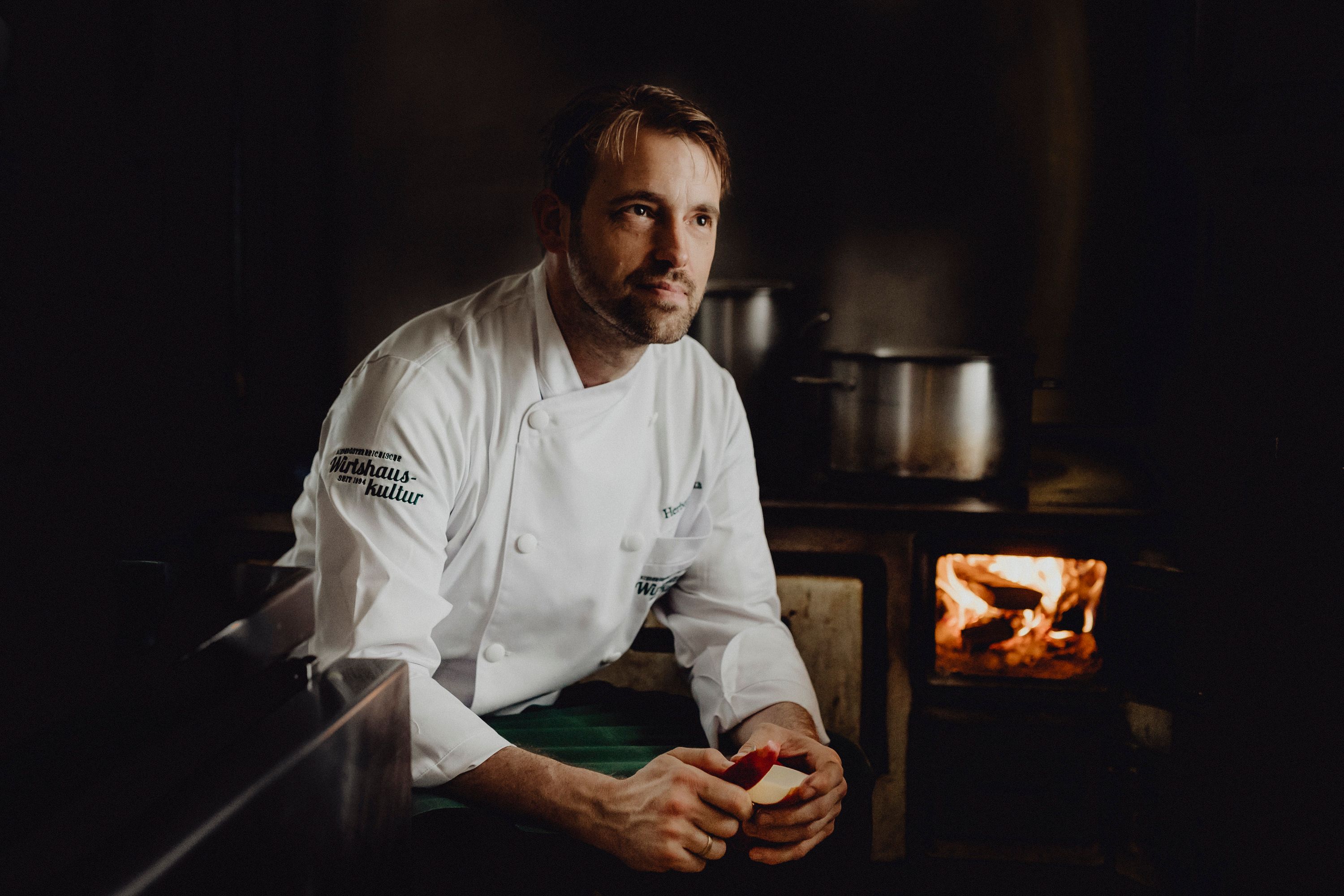 Koch in weißer Uniform vor einem traditionellen Herd mit Feuer.