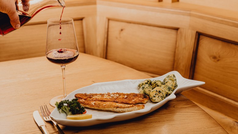 Ein Teller mit Zanderfilet und Kartoffeln in Knoblauchbutter, daneben ein Glas Rotwein auf einem Holztisch.