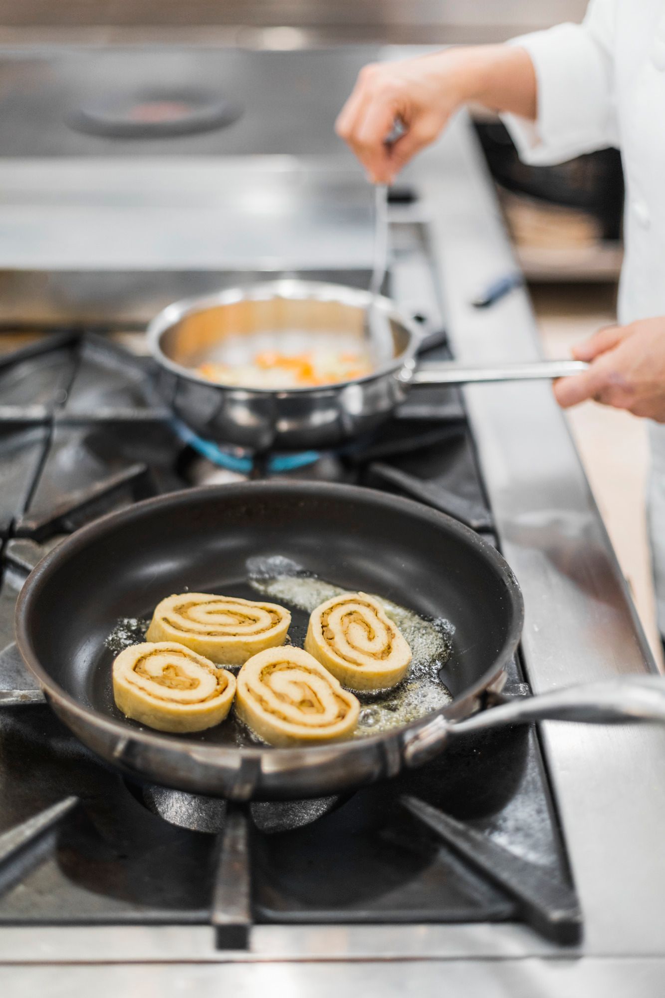 Eine Pfanne mit drei Steinpilz-Rouladen auf einem Herd, daneben eine Hand, die eine andere Pfanne hält.