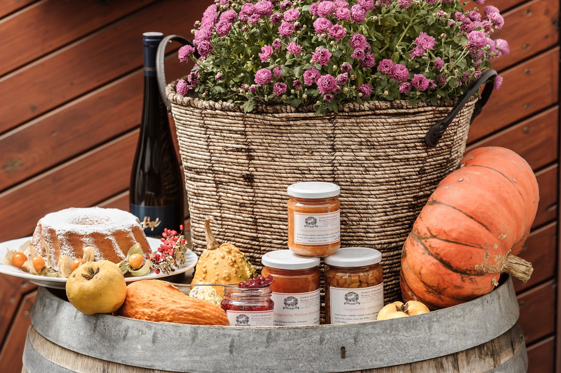 Ein Arrangement aus hausgemachten Spezialitäten mit Kürbis, Marmelade, Kuchen und Blumen in einem Korb auf einem Fass.