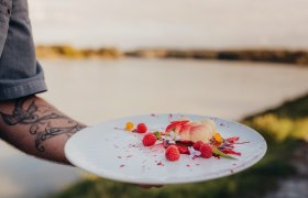 Ein Teller mit wei&szlig;em Schokomousse, Himbeeren und Bl&uuml;ten, gehalten von einer t&auml;towierten Hand vor einem See.