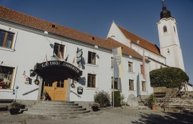 Ein Gasthof mit der Aufschrift 'Gasthof Gamerith' neben einer Kirche mit Turm unter blauem Himmel.