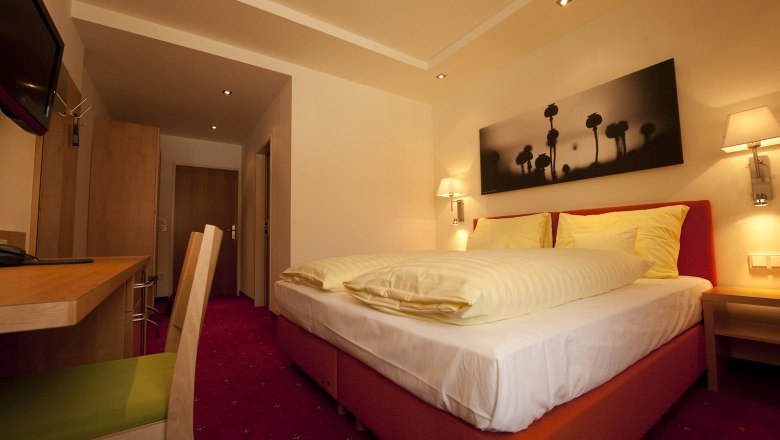 Ein gemütliches Hotelzimmer mit Doppelbett, Schreibtisch und Wandbild. Warme Beleuchtung und rote Teppiche schaffen eine einladende Atmosphäre.