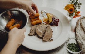 Ein Teller mit Fleischscheiben, Kroketten, Preiselbeeren und Zitronenscheiben wird angerichtet. Daneben steht ein kleines Blumenarrangement.