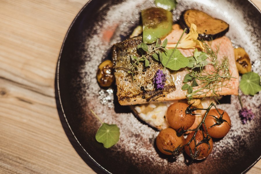 Lachsforellenfilet auf Grammelrisotto mit Gem&uuml;se und Kr&auml;utern auf einem Teller.