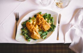 Ein Teller mit gebackenem Karpfenfilet auf Feldsalat und Kartoffeln, auf einem weiß gedeckten Tisch.