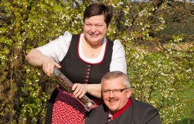 Ein Mann und eine Frau in traditioneller Kleidung lächeln im Freien, die Frau hält eine große Pfeffermühle.