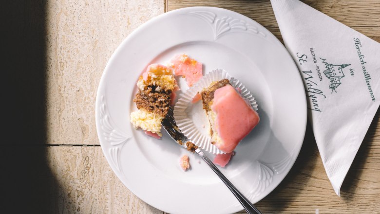 Ein Punschkrapferl auf einem Teller mit einer Gabel, daneben eine Tasse Kaffee und ein Glas Wasser auf einem Tisch.