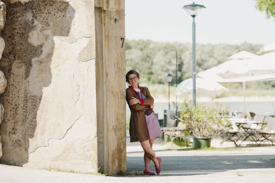Frau lehnt l&auml;chelnd an einer Mauer, im Hintergrund Terrasse mit Tischen, St&uuml;hlen und Sonnenschirmen.
