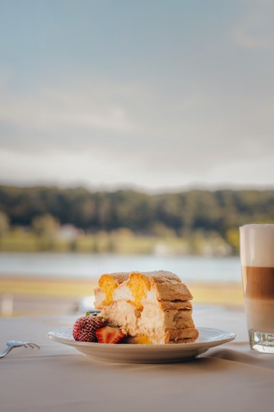 Kuchenstück mit Erdbeeren und Latte Macchiato vor unscharfer Landschaft.