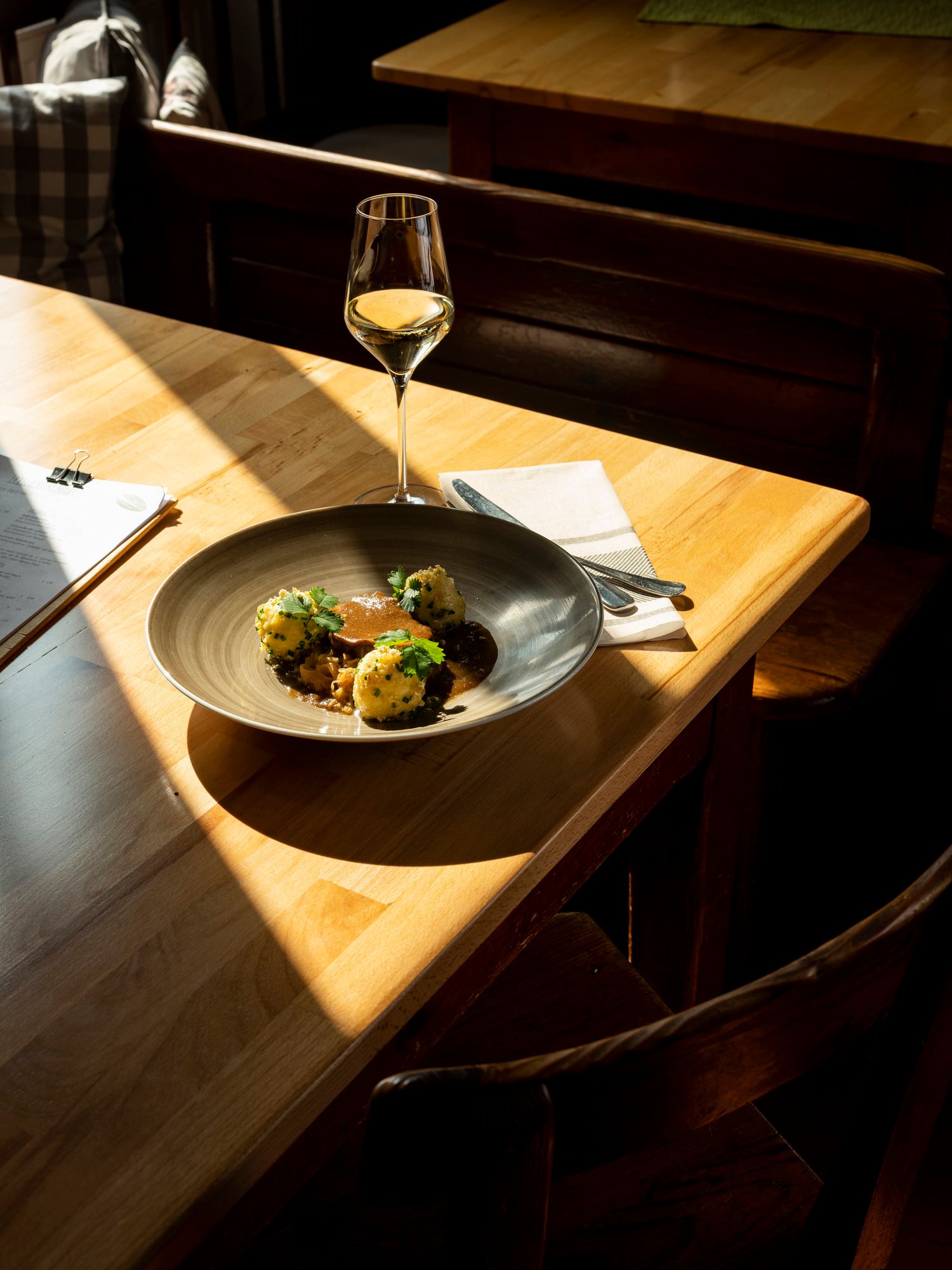 Teller mit Fleisch, Knödeln und Soße, Weinglas auf Holztisch im Sonnenlicht.