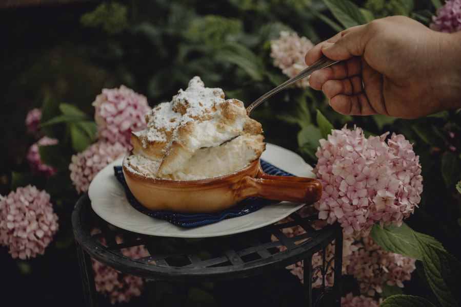 Ein Souffl&eacute; in einer braunen Schale, umgeben von rosa Hortensien, wird mit einem L&ouml;ffel serviert.