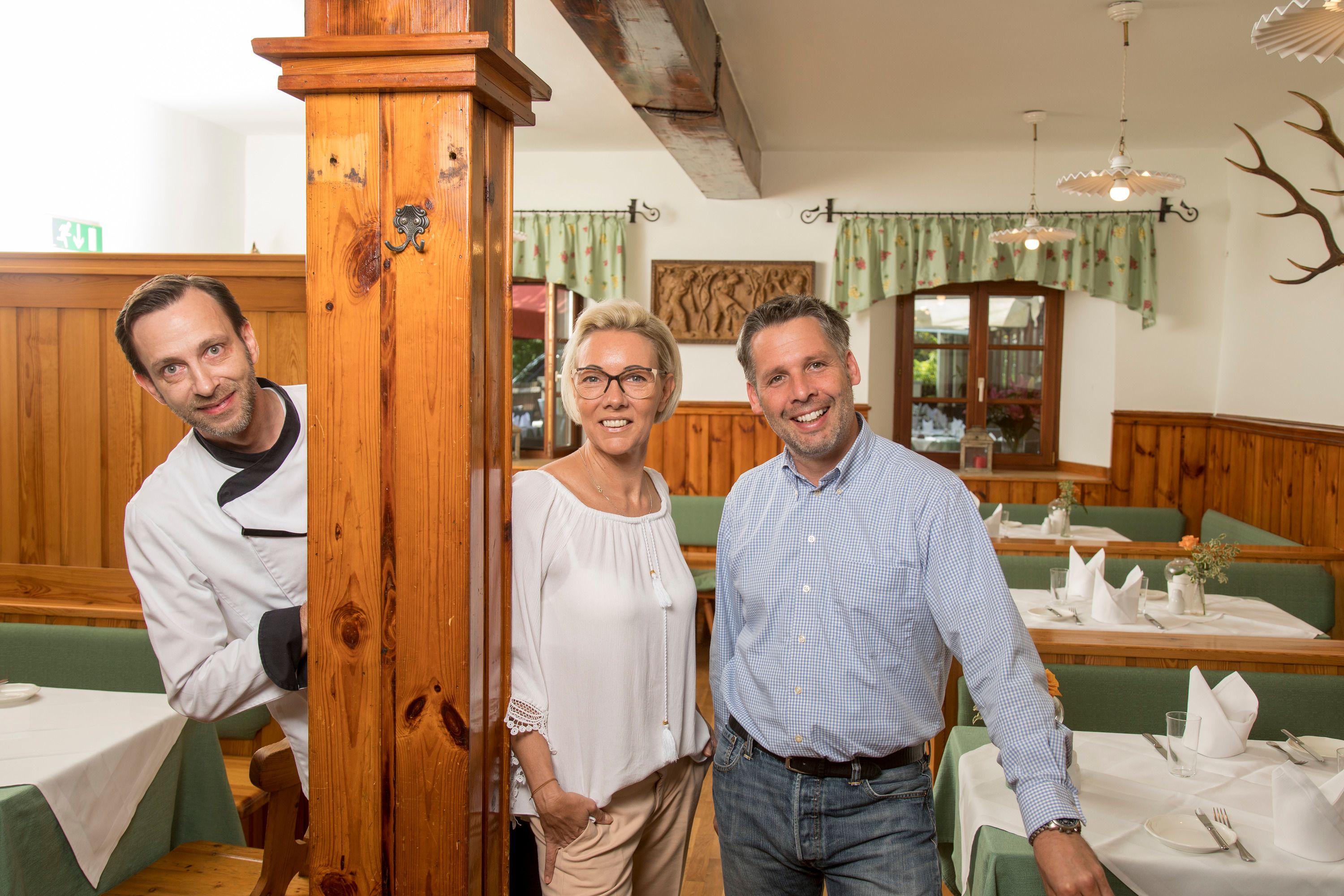 Drei Personen in einem rustikalen Restaurant, eine Person in Kochkleidung, zwei in Freizeitkleidung, lächeln in die Kamera.