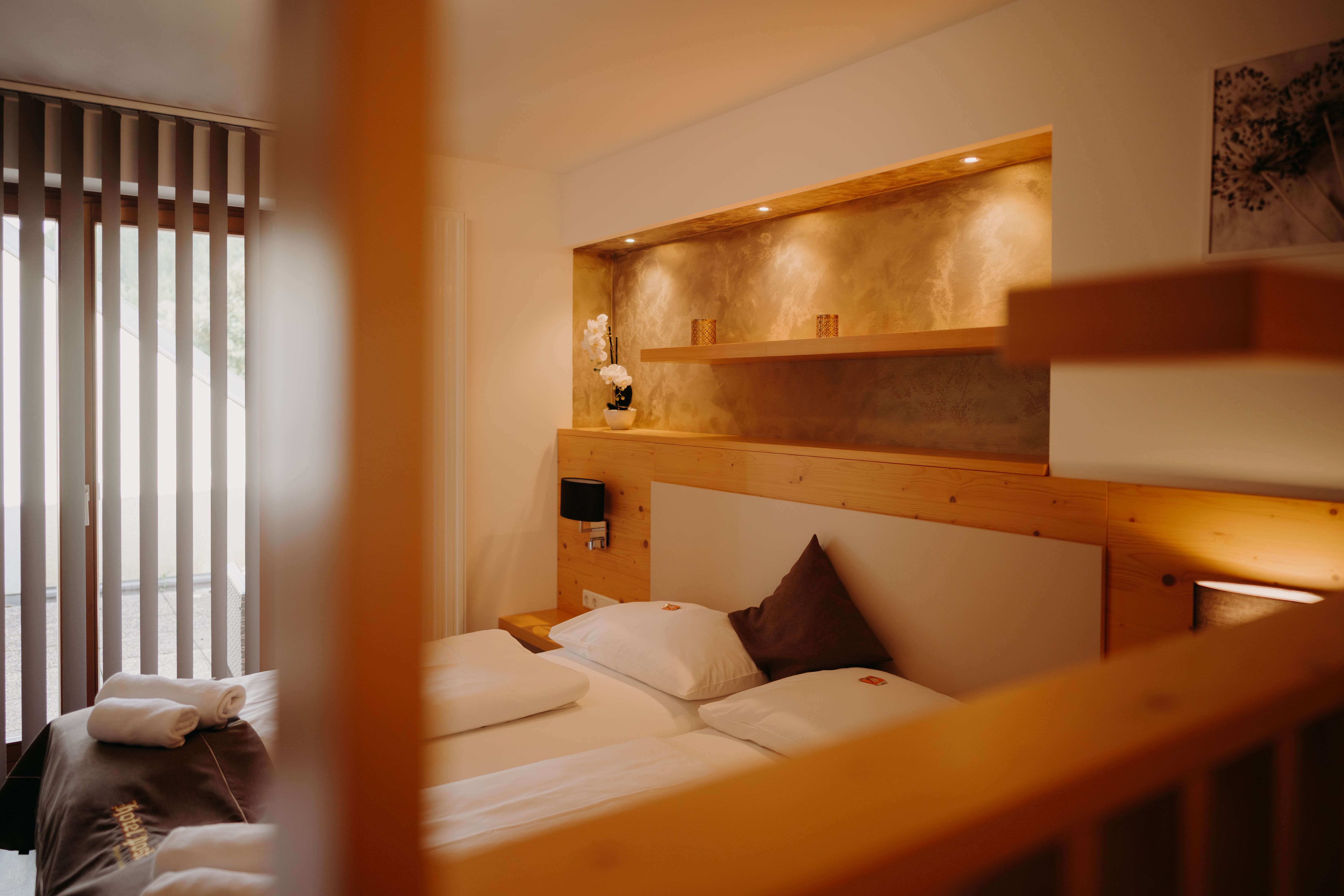 Gemütliches Appartement mit Doppelbett und Balkontür. Die Wand ist zum Teil mit hellem Holz verkleidet, hinter dem Bett ist eine in die Wand eingelassene Nische mit Ablagefläche und Spots, in einem erdigen Gold gestrichen.