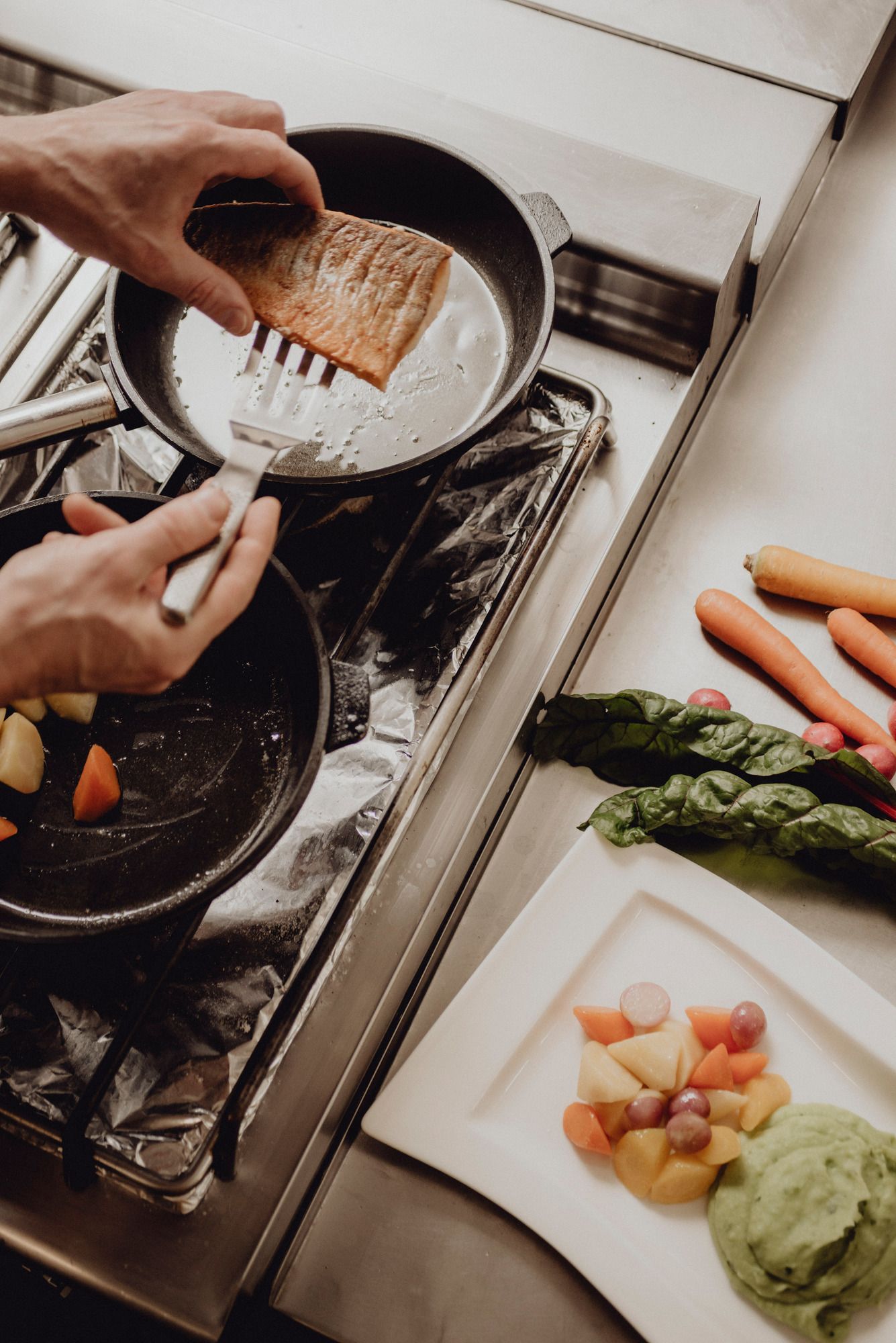 Eine Person brät ein Stück Fisch in einer Pfanne auf einem Herd. Daneben liegen Gemüse und ein Teller mit Beilagen.