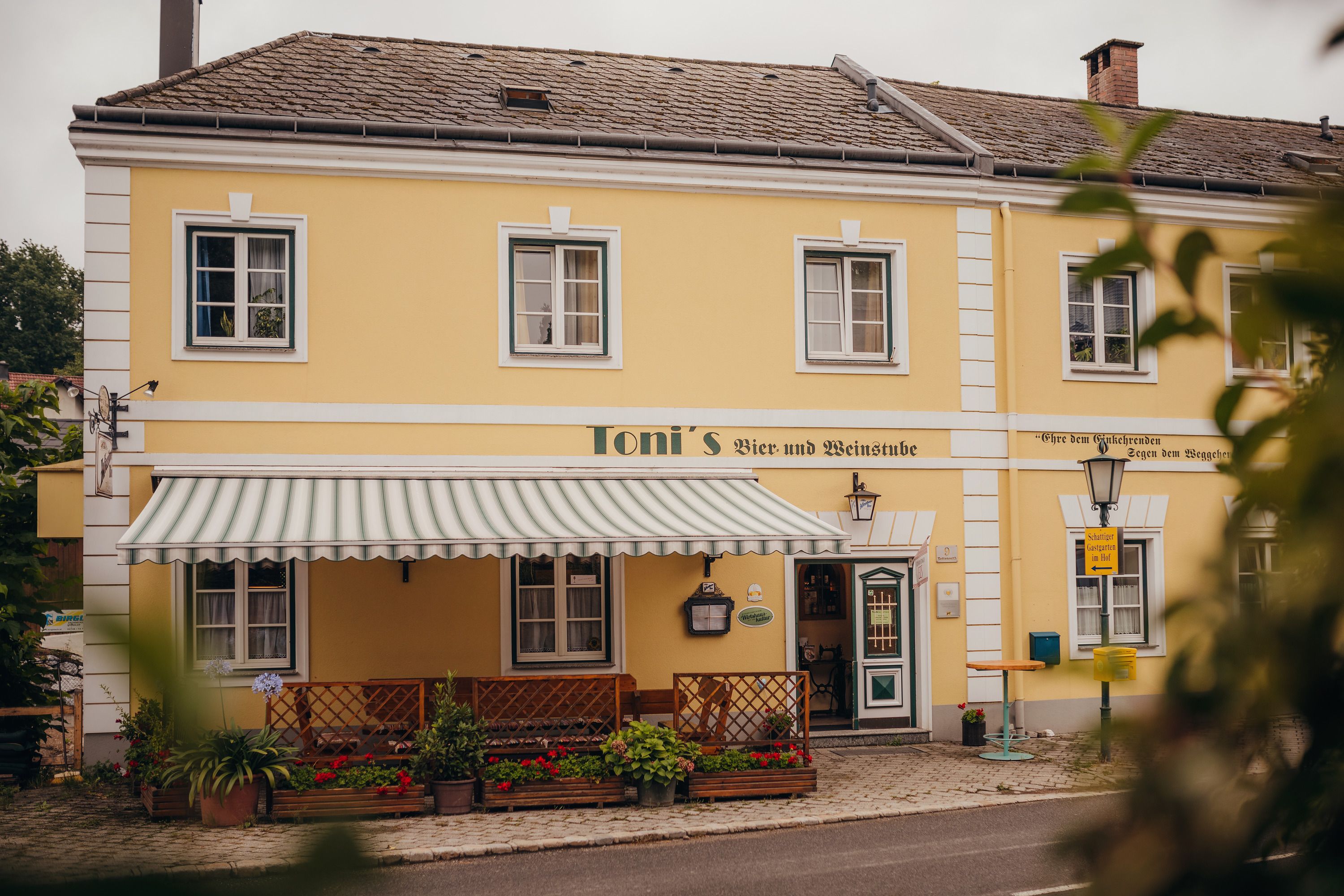 Yellow building with awning and plants, labeled 'Toni's Bier- und Weinstube'.