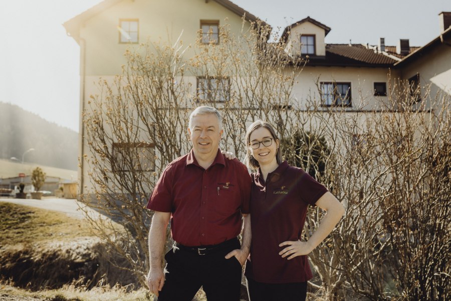 Zwei Personen in roten Hemden stehen l&auml;chelnd vor einem Geb&auml;ude.