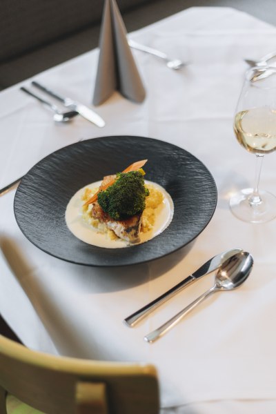 Ein Teller mit Zander auf Erd&auml;pfelp&uuml;ree, garniert mit Brokkoli und Karotten, auf einem gedeckten Tisch mit Besteck und einem Glas Wei&szlig;wein.