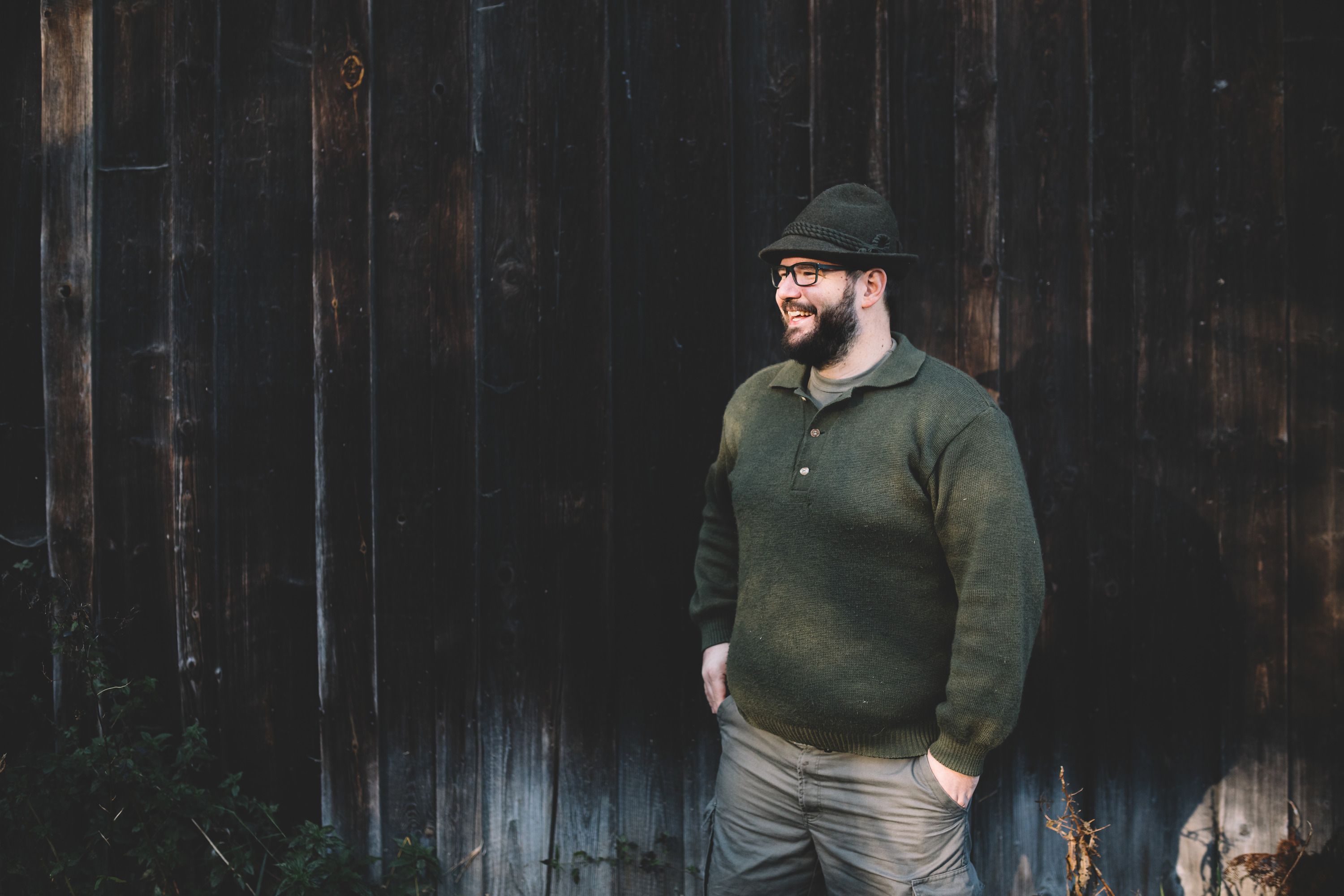 Mann in grüner Kleidung und Hut steht vor einer Holzwand.