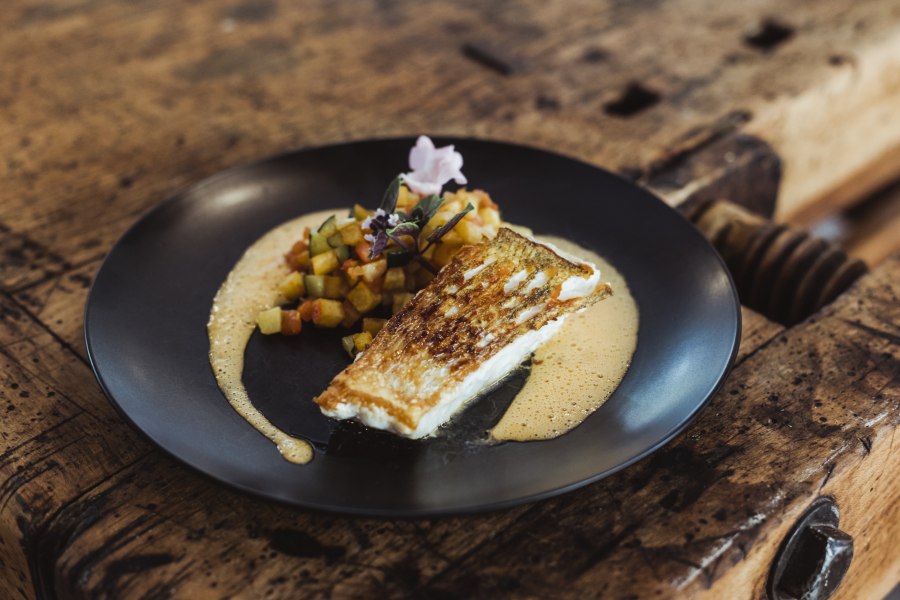 Gebratener Fisch mit Gem&uuml;sew&uuml;rfeln und Sauce auf schwarzem Teller serviert.