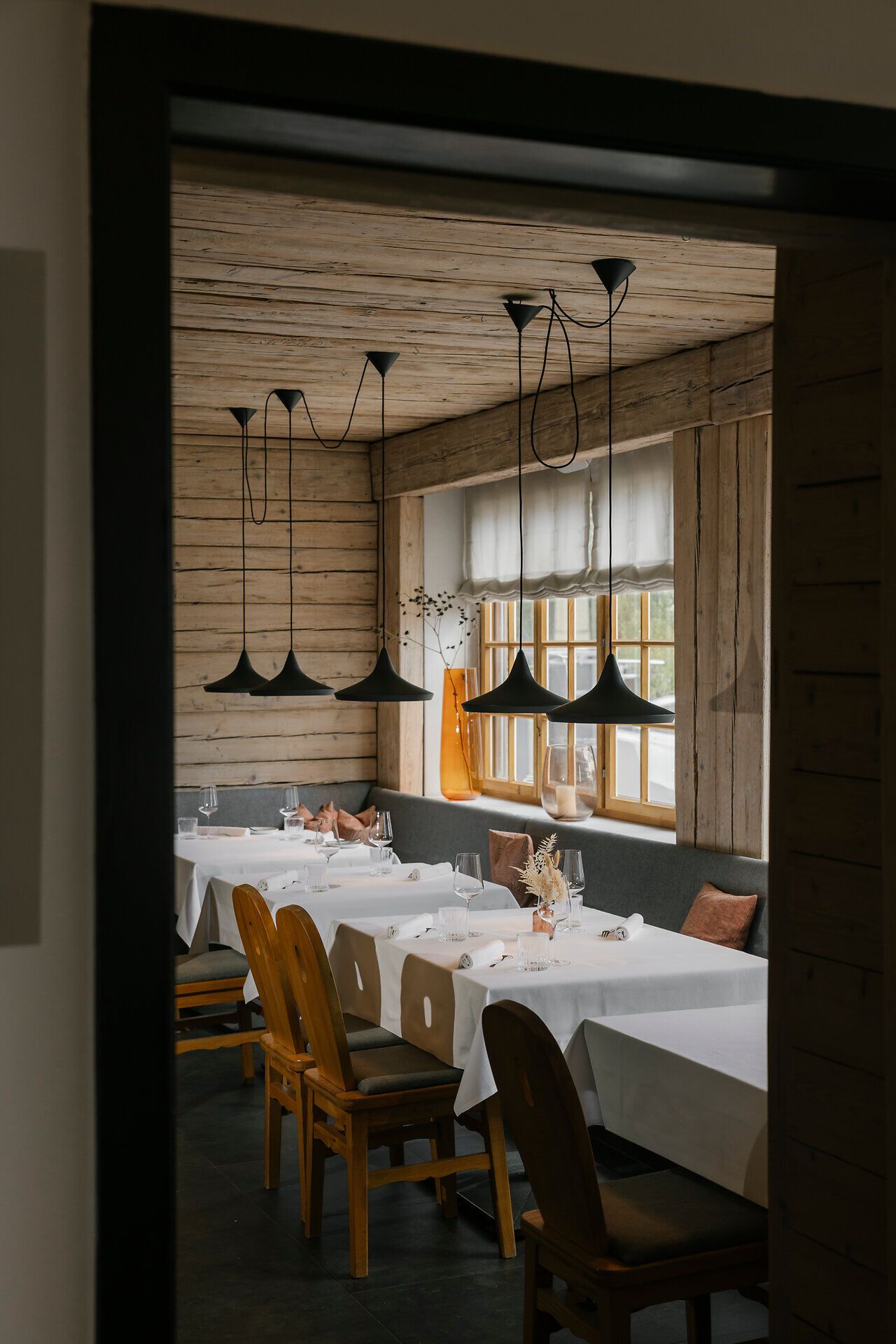 Gastraum mit Holzwänden, gedeckten Tischen, Holzstühlen und Sitzbank. Schwarze Pendelleuchten hängen über den Tischen, Tageslicht fällt durch die Fenster.